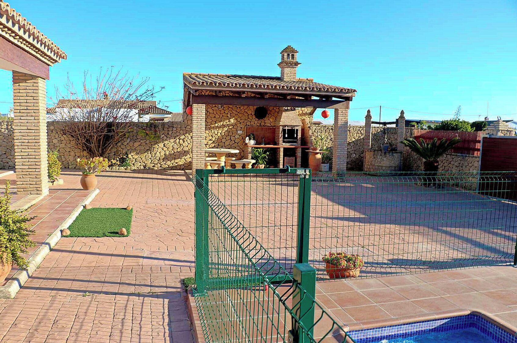 A backyard with a swimming pool, a stone patio, and a small brick structure with a tiled roof.