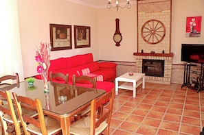 A living room with a red sofa, wooden dining table, chairs, a fireplace, and a TV. - Rustic holiday villa for 5 people (CONIL DE LA FRONTERA)