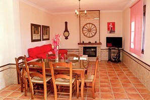 A dining area with a glass table and wooden chairs, a red sofa, a fireplace, and a clock on the wall. - Rustic holiday villa for 5 people (CONIL DE LA FRONTERA)