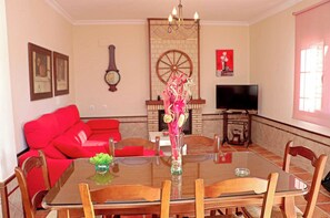 A dining area with a glass table, wooden chairs, a red sofa, a decorative vase, and a television in the background.