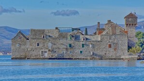 A stone building with multiple windows and a tower, situated by a body of water with mountains in the background. - Modern apartment near Split and Trogir (Kaštel Gomilica)