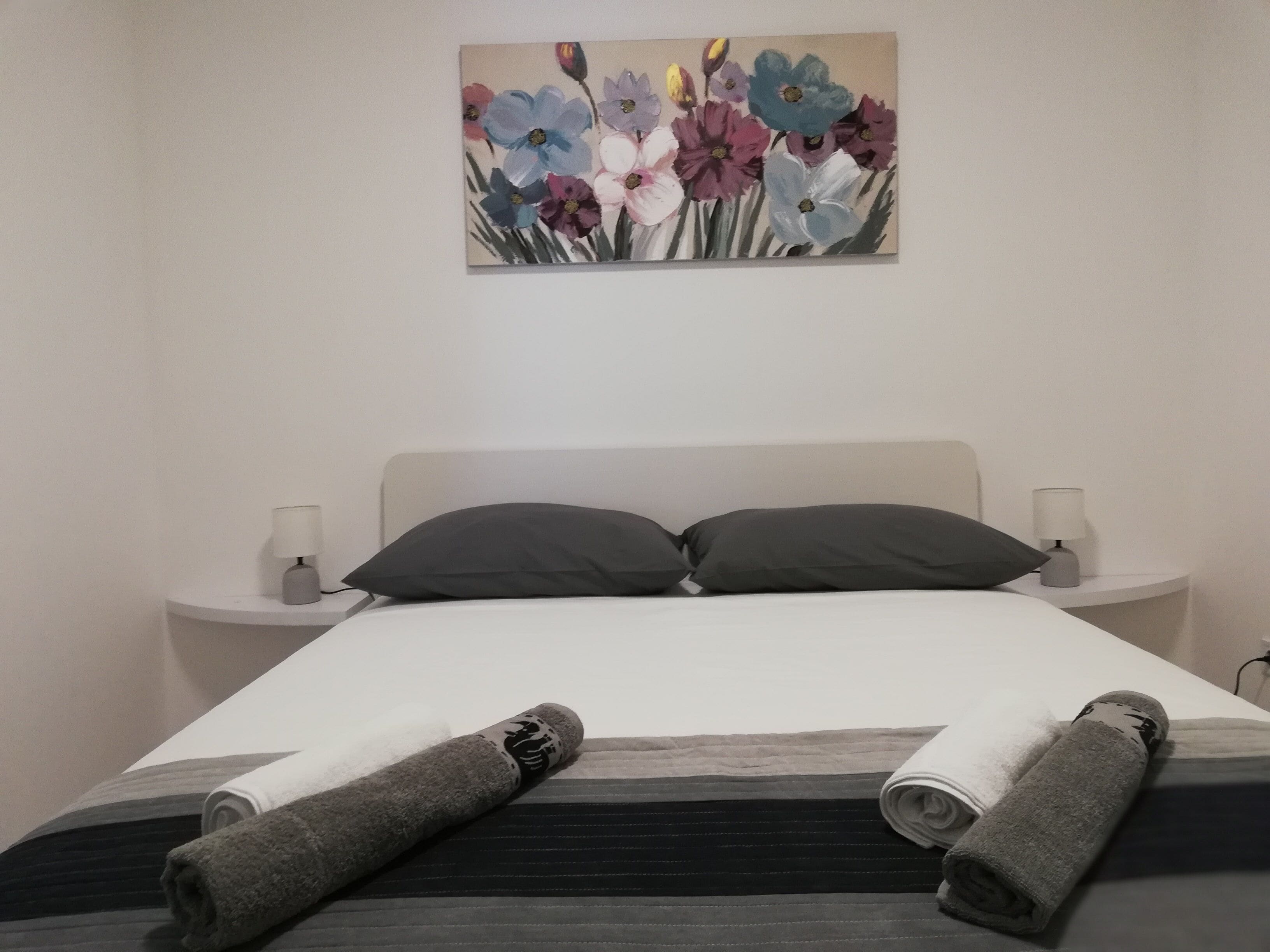 A neatly made bed with dark bedding, two pillows, and two rolled towels. There is a painting of flowers on the wall above the bed.