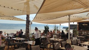 An outdoor dining area with tables and chairs, palm trees, and a view of the sea. - Modern apartment near Split and Trogir (Kaštel Gomilica)