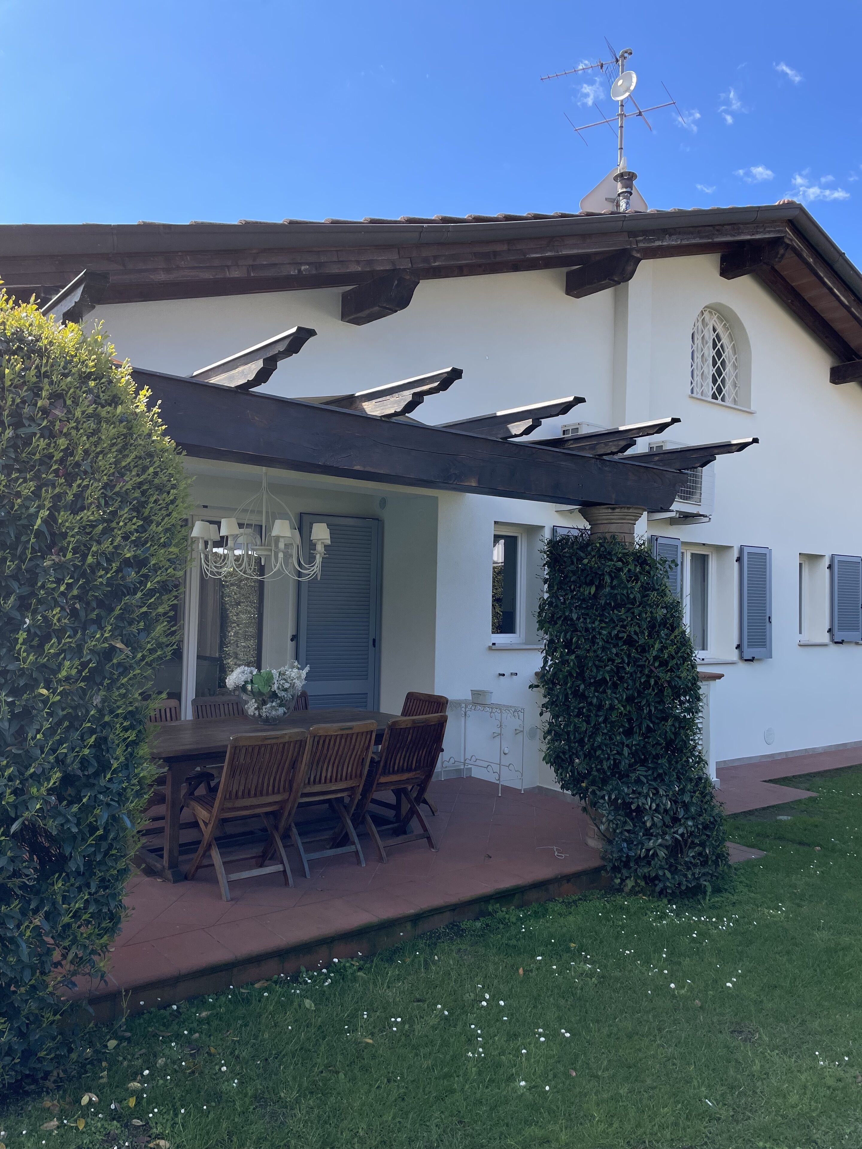 Une terrasse couverte avec une table et des chaises, un petit jardin et un bâtiment à la façade blanche et aux volets.