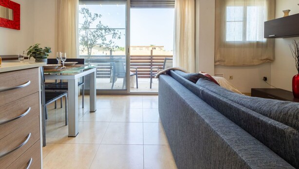 A modern living room with a sofa, dining table, and kitchen area. There is a sliding glass door leading to an outdoor balcony.