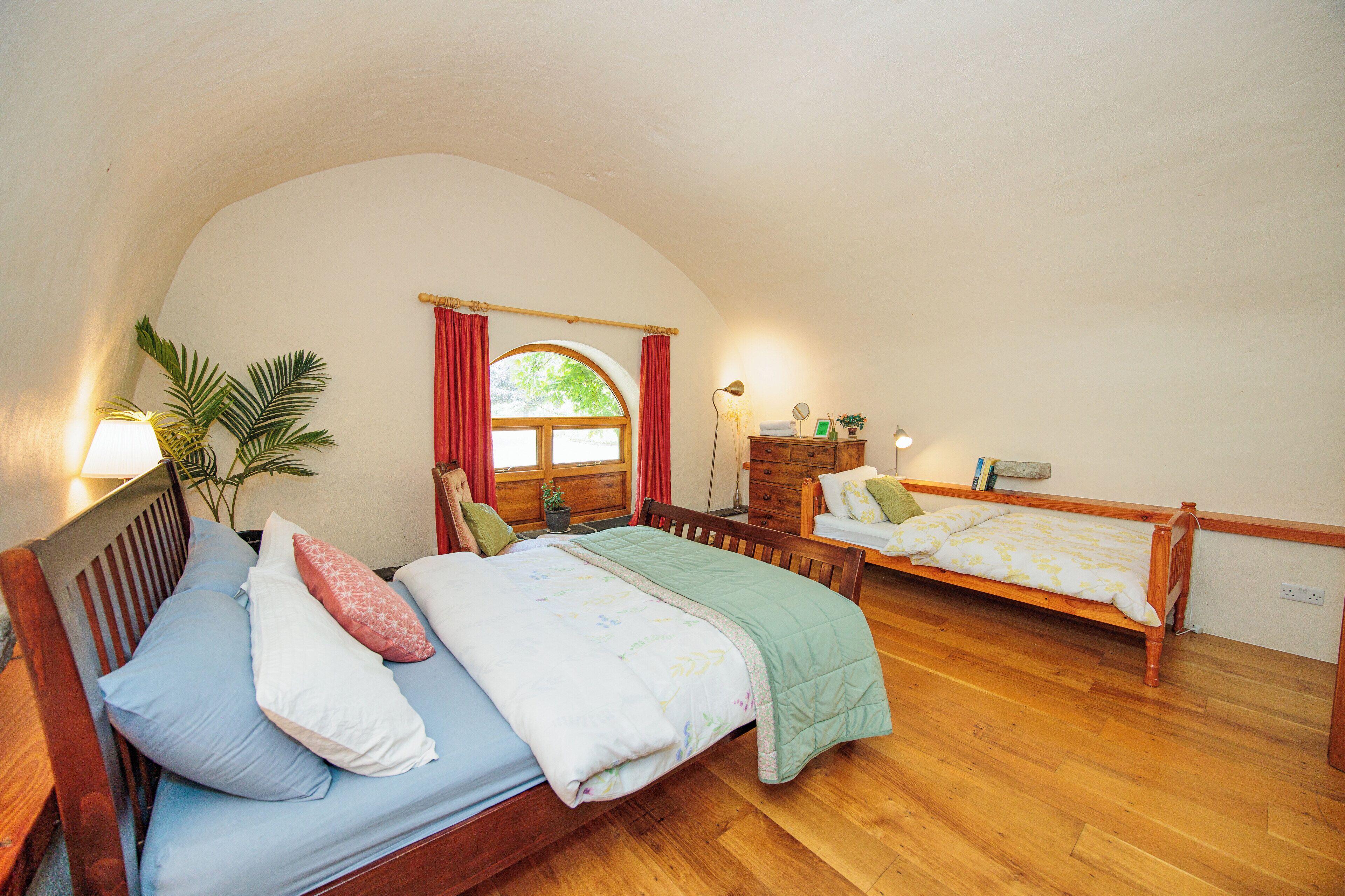 A cozy bedroom with two wooden beds, a large window with red curtains, a bedside table with a lamp, and a potted plant.