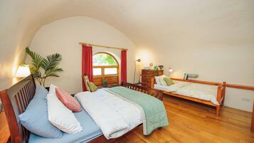 A cozy bedroom with two wooden beds, a large window with red curtains, a bedside table with a lamp, and a potted plant.