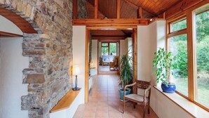 A hallway with a stone wall, wooden ceiling beams, a tiled floor, a chair, a table, and a potted plant by a window. - 18th Century Coachhouse in a Courtyard Setting (Inistioge)