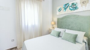 Una cama bien hecha con cubrecama blanco y almohadas estampadas en verde, una mesita de noche con lámpara y una pared con un cuadro floral.