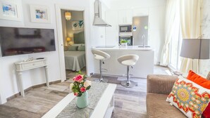 Un salón moderno con televisor de pantalla plana, una mesa baja con flores, un sofá con un cojín colorido y una zona de cocina con microondas y fregadero.