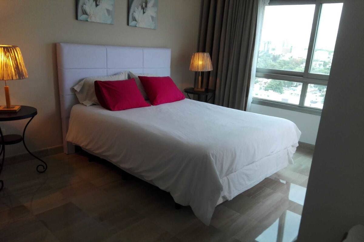 A hotel room with a large bed, two bedside lamps, a view of the city through the window, and artwork on the wall.