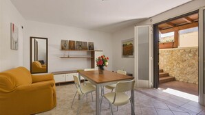 A modern living room with a wooden dining table, two chairs, a sofa, and a coffee table with flowers. There is a mirror, artwork on the wall, and a view of the outside through a glass door. - Girasole Trilo 2 by BarbarHouse (Castrignano del Capo)