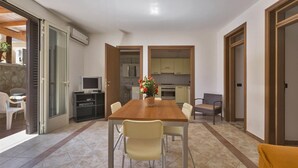 A dining area with a wooden table and chairs, a TV on a stand, and a kitchen in the background. - Girasole Trilo 2 by BarbarHouse (Castrignano del Capo)