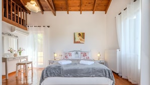Quarto com piso de madeira, cama com manta cinza, cabeceira branca, quadro acima da cama, escrivaninha com luminária e janela com cortinas leves.