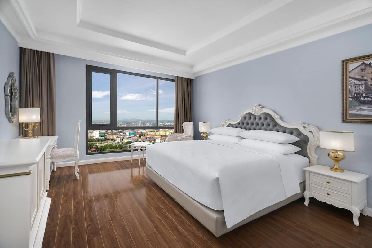 Une chambre avec un grand lit, une table de chevet, une chaise et une vue sur le paysage urbain, visible à travers une grande fenêtre.