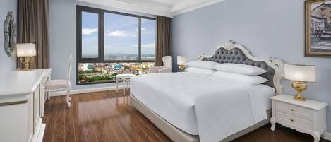 Un dormitorio con una cama grande, una mesita de noche, una silla y vistas a un paisaje urbano a través de un ventanal.