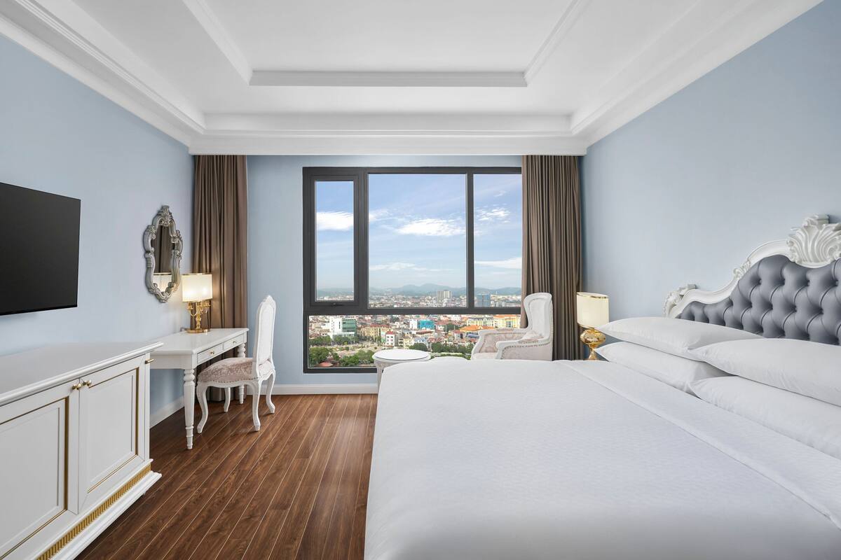 Une chambre d’hôtel avec un grand lit, un bureau, une chaise et une vue sur la ville grâce à de grandes fenêtres.