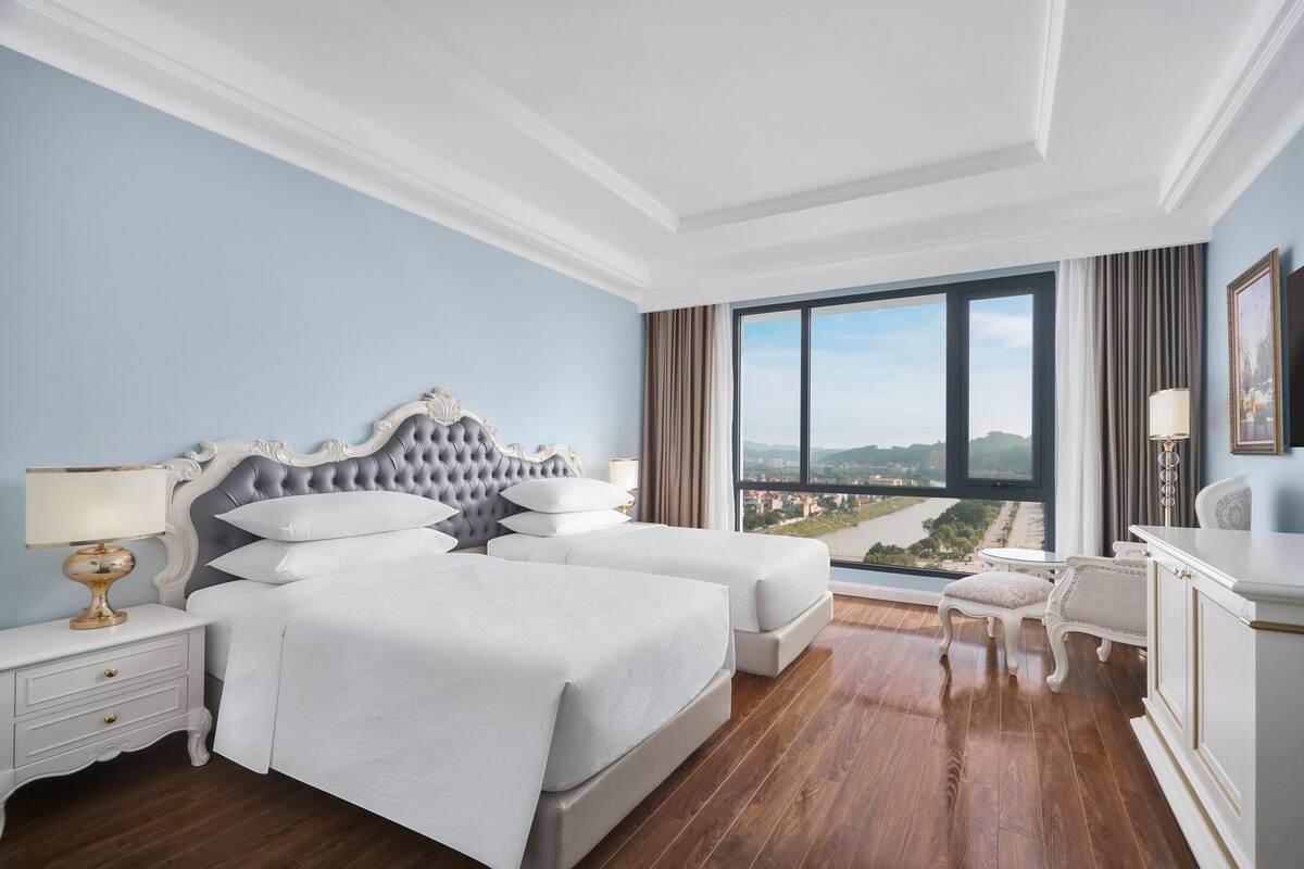 Une chambre d’hôtel avec un grand lit, deux tables de chevet, un bureau avec une chaise et une vue sur le paysage urbain par une grande fenêtre.