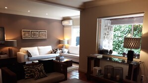A living room with a sofa, coffee table, and a TV stand. There is a window with a view of greenery outside.