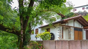 A rustic house with a stone facade, wooden beams, and a garden with flowers and a tree.