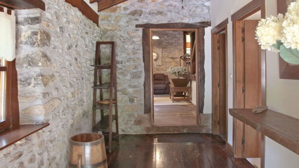 A stone-walled hallway with wooden beams, a wooden barrel, and a view into a cozy living area with a sofa and a table.