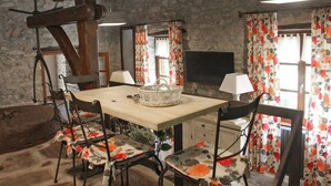A rustic dining room with a stone wall, wooden beams, a wooden table, floral chairs, a chandelier, and a fireplace.