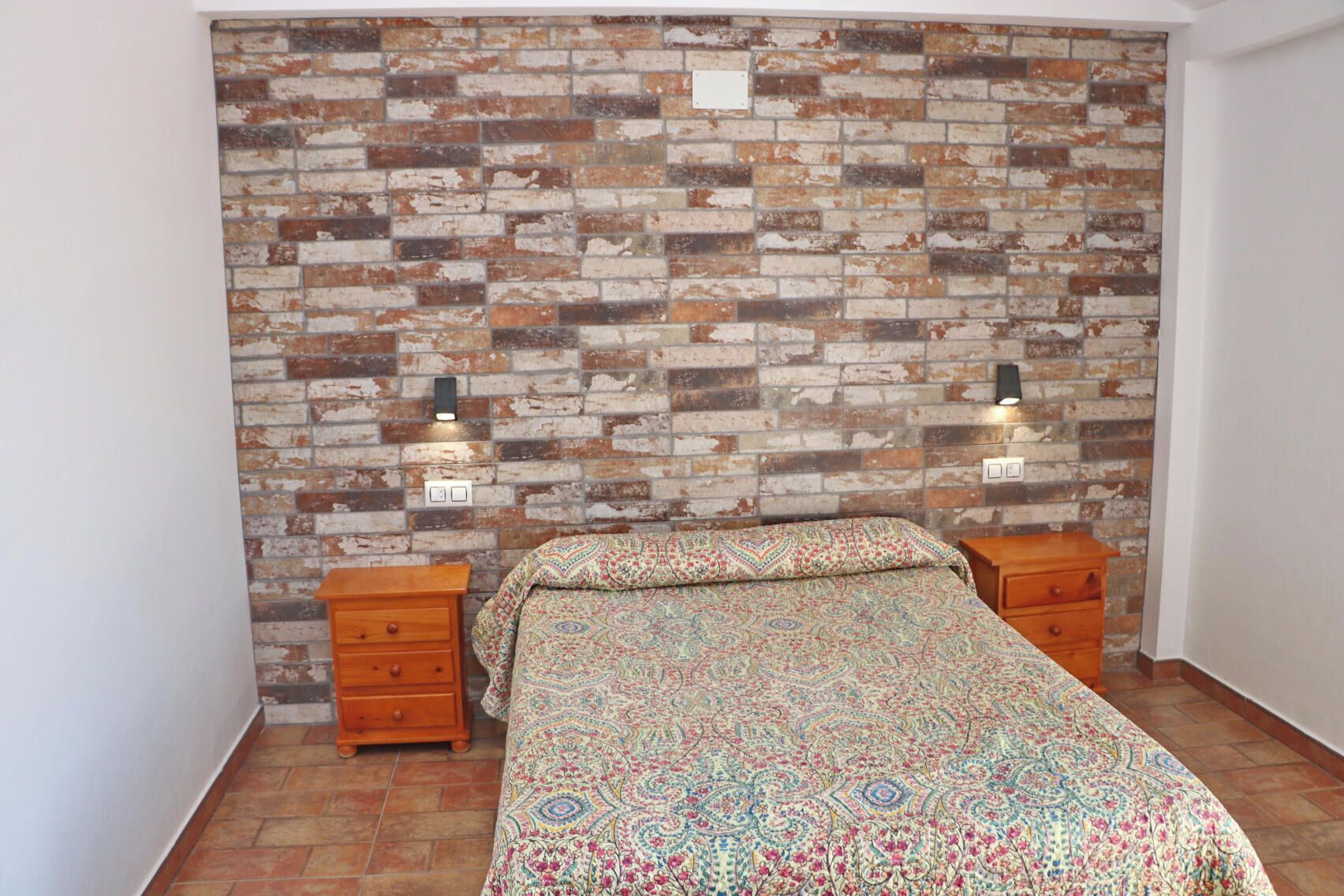 Une chambre à coucher avec une tête de lit ornée d’un motif en briques, une couvre-lit à motifs et deux tables de chevet équipées de lampes murales.