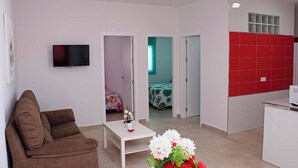 Una sala de estar moderna con un sofá marrón, una mesa de centro blanca y un televisor montado en la pared. Se ve un acceso a un dormitorio con cama y un baño con armarios rojos.