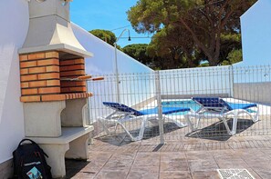 Un área de barbacoa con horno de ladrillo, una piscina con sillas de descanso y un árbol al fondo.