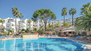 Piscina con sillas de descanso y sombrillas, rodeada de palmeras y un edificio con balcones.