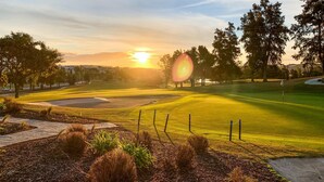 Un campo de golf al atardecer con cielo despejado, árboles y una vista distante de casas.