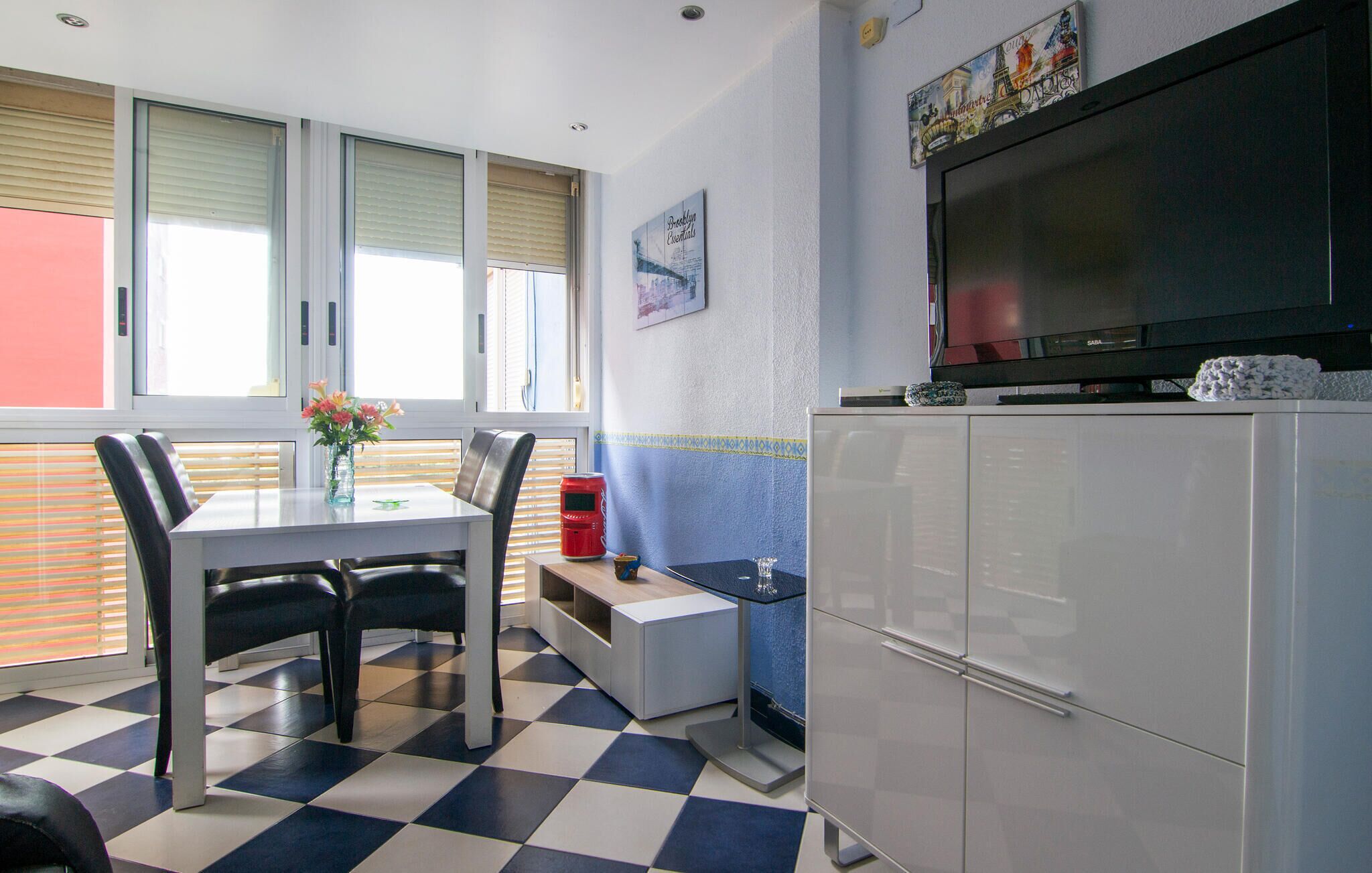 A compact kitchen with a dining area, featuring a checkered floor, a refrigerator, a television, and a window with blinds.