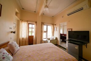A bedroom with a bed, a television, a refrigerator, a window with curtains, and a door leading to a bathroom.