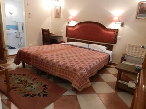 A hotel room with a bed, a desk, a chair, a small table, a rug, and a shower area. - Hotel Arya Niwas, Jaipur (Jaipur)