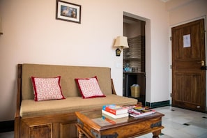 A living room with a sofa, two pillows, a coffee table with books, a lamp, and a wooden door. - Hotel Arya Niwas, Jaipur (Jaipur)