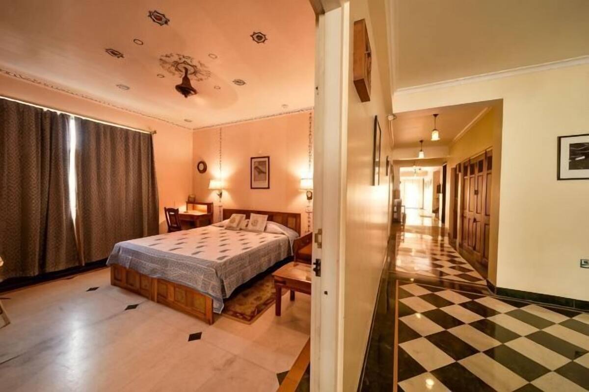 Ein Hotelzimmer mit Bett, Nachttischen, einem Schreibtisch, einem Stuhl, einem Fenster mit Vorhängen, einem Korridor mit Schachbrettmuster und einer Tür zu einem anderen Raum.