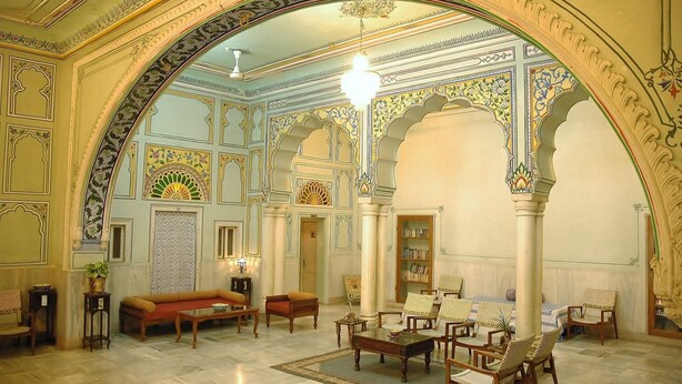 An ornate room with a high ceiling, intricate wall designs, a sofa, chairs, a table, and a bookshelf. - Hotel Arya Niwas, Jaipur (Jaipur)