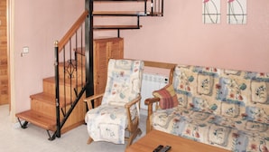 Una sala de estar con una escalera de madera, un sofá, una silla y un control remoto sobre una mesa.