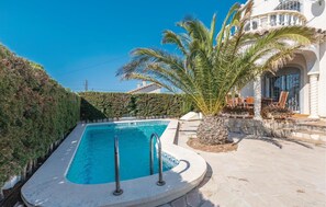 A rectangular swimming pool with a stone deck, surrounded by a hedge and a palm tree.