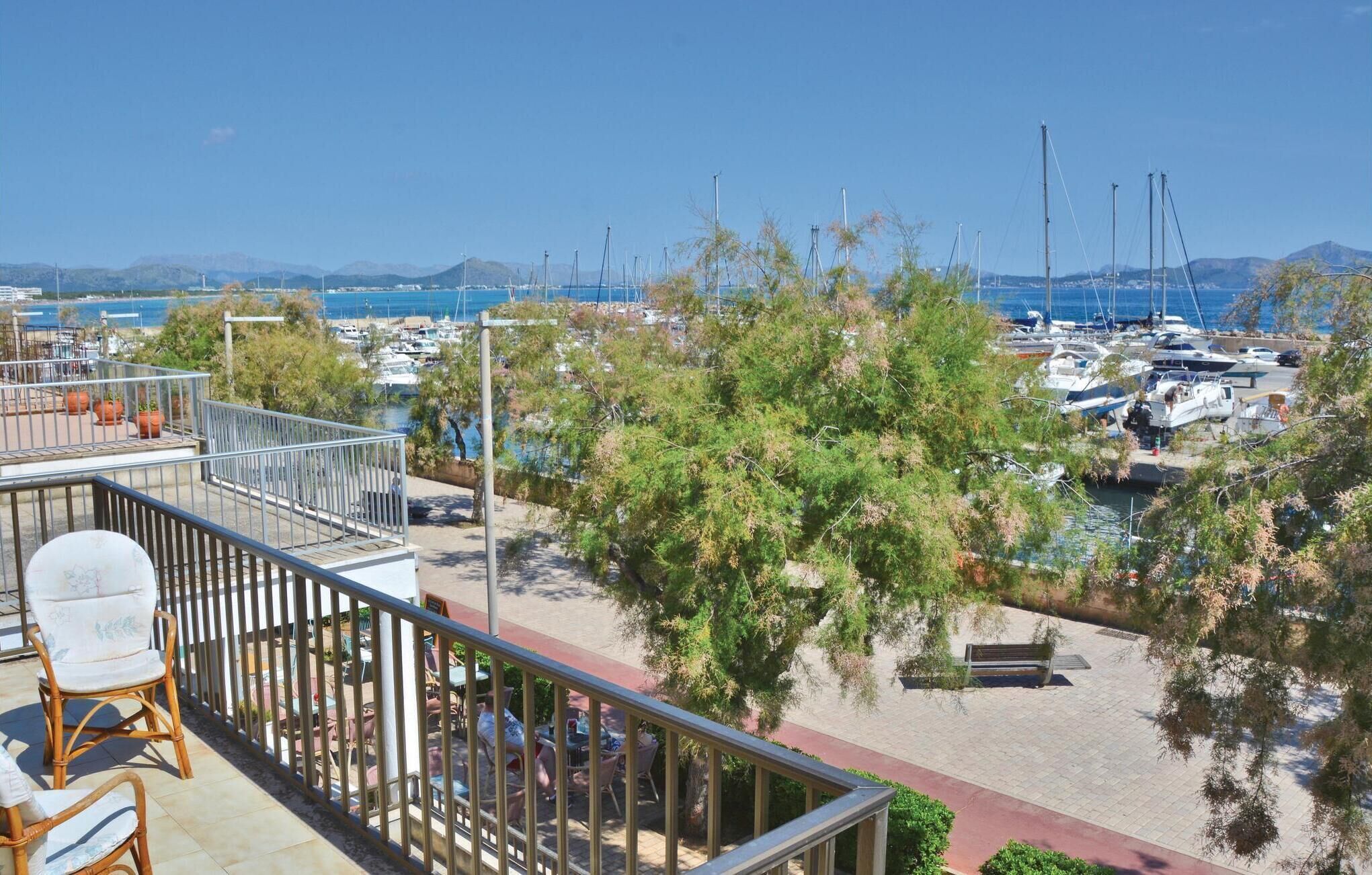 Un balcone con sedie in vimini e un tavolo, affaccia su un porto turistico con barche e una vista limpida sul mare e montagne in lontananza.