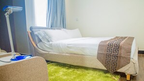 A neatly made bed with a patterned throw, a standing lamp, and a green carpet. - PCG Gisimenti Apartment (Kigali)
