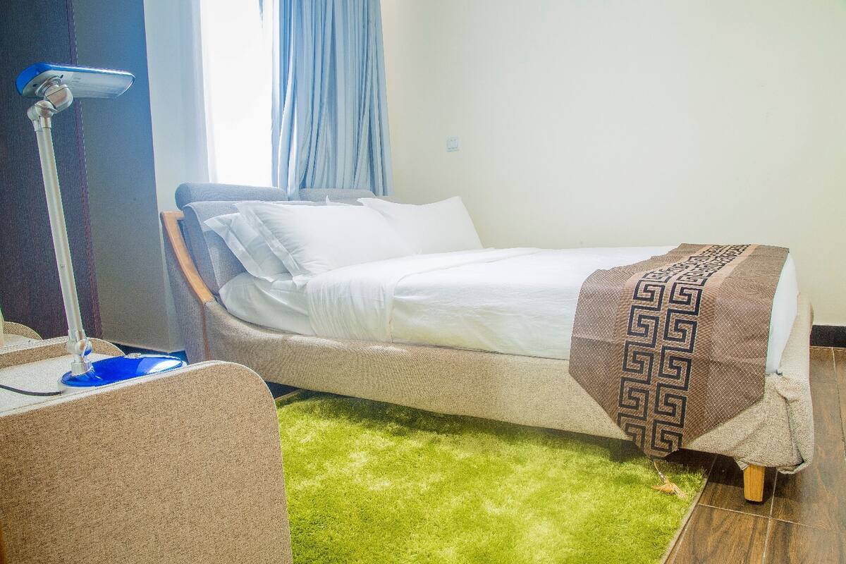 Un lit bien fait, recouvert d’un couvre-lit à motifs, une lampe sur pied et une moquette verte.