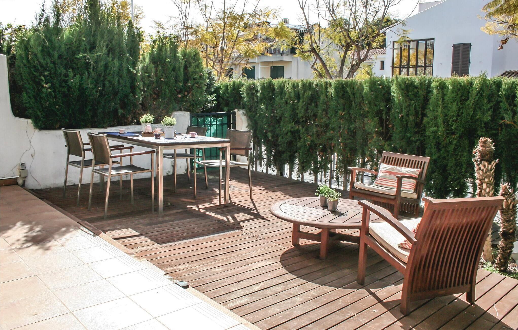 Een houten terras met een tafel en stoelen, omgeven door groen en een wit gebouw op de achtergrond.