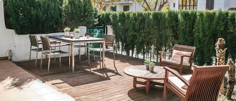 Een houten terras met een tafel en stoelen, omgeven door groen en een wit gebouw op de achtergrond.