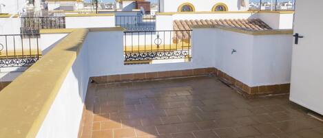 Una terraza en la azotea con suelo de baldosas, una pequeña ventana con rejas de hierro decorativas y una barandilla amarilla.