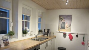 Eine moderne Küche mit weißen Schränken, einer Holz Arbeitsplatte, einem Geschirrspüler, einer Spüle und einem Mülleimer. Auf der Fensterbank stehen Blumentöpfe und an der Wand hängt ein Gemälde.
