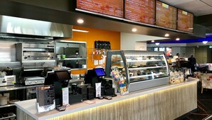 Um balcão de restaurante com um quadro de menu acima, uma estação de autoatendimento para bebidas e uma vitrine com sobremesas.