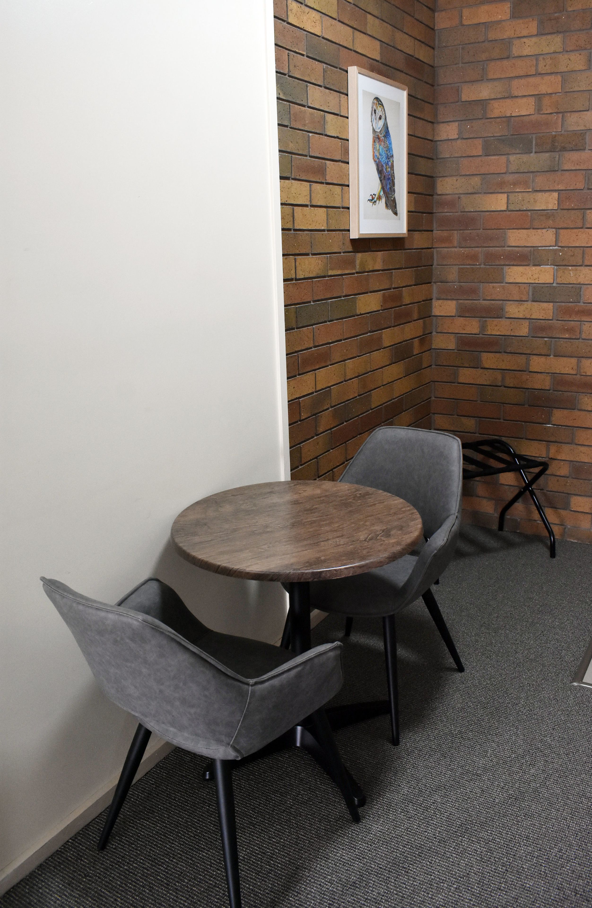 A corner office with a round wooden table, two grey upholstered chairs, a black metal chair, and a framed owl artwork on a brick wall.