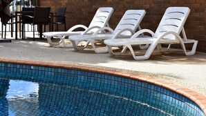 Uma área ao lado da piscina, com espreguiçadeiras brancas e um muro de tijolos.