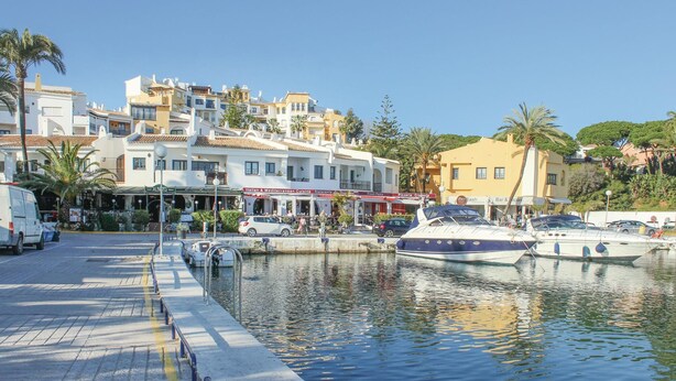 Una marina con barche ormeggiate, una passeggiata sul lungomare e edifici con negozi e ristoranti.